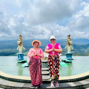 Two women in traditional attire posing by a scenic fountain.